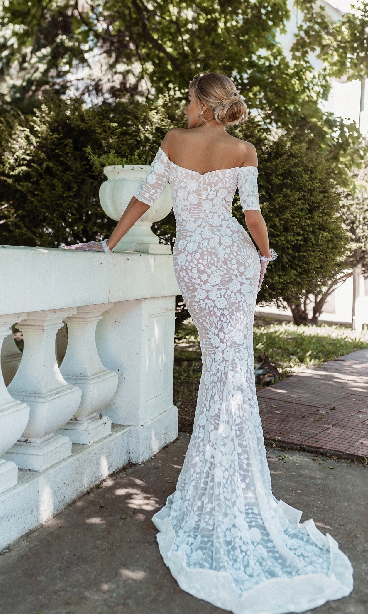 A woman stands outdoors by a white railing, facing away in the Off-Shoulder White Bridal Gown: Tina Holly BB009, its beaded lace and long mermaid silhouette flowing gracefully.